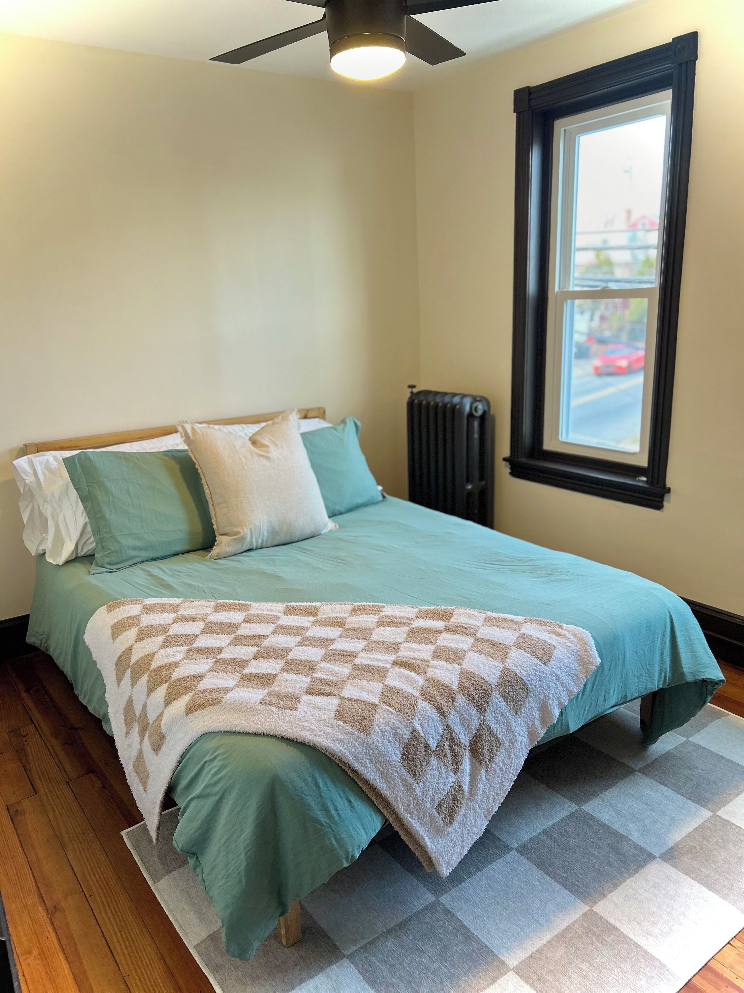 Une chambre à coucher avec un lit, des oreillers, un radiateur, une fenêtre et un ventilateur de plafond.