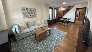 Ein Wohnzimmer mit grauem Sofa, einem Holztisch und einem gemusterten Teppich. Eine Treppe führt zu einer oberen Ebene.