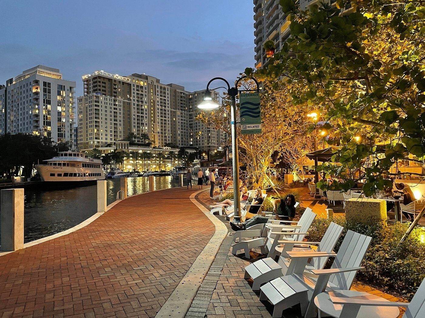 A waterfront promenade with outdoor seating, a river, and high-rise buildings in the background.