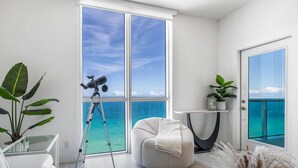 A modern living room with a large window overlooking a turquoise sea, a telescope on a tripod, a comfortable sofa, and a small table with plants. - Beachfront Luxe Corner Penthouse|Gym|Heated Pool (Hollywood)