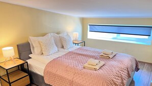 Een netjes opgemaakt bed met een roze sprei, twee bedlampjes en een raam met jaloezieën.