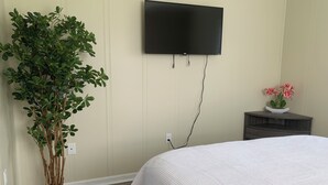 Un dormitorio con una cama, una planta en maceta, un televisor montado en la pared y una mesita de noche con un florero.