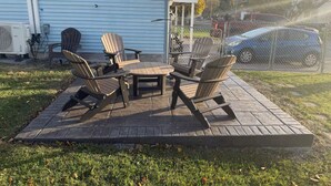 Un patio con sillas Adirondack y una mesa, una casa azul al fondo y un coche aparcado detrás de una valla.