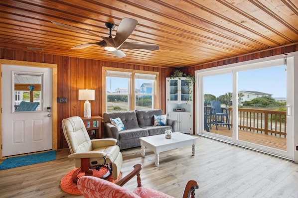 A spacious living room with a ceiling fan, a sofa, a coffee table, and a view through large sliding glass doors to an outdoor deck.