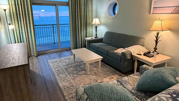 A living room with a sofa, a coffee table, a side table with a lamp, and a view of the ocean through a balcony.