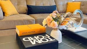 A living room with a brown leather sofa, a black coffee table with a Louis Vuitton box and a vase of flowers, and a wall decoration above the sofa.