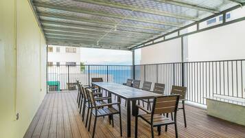 A covered outdoor area with a long table and chairs, a railing, and a view of the sea.
