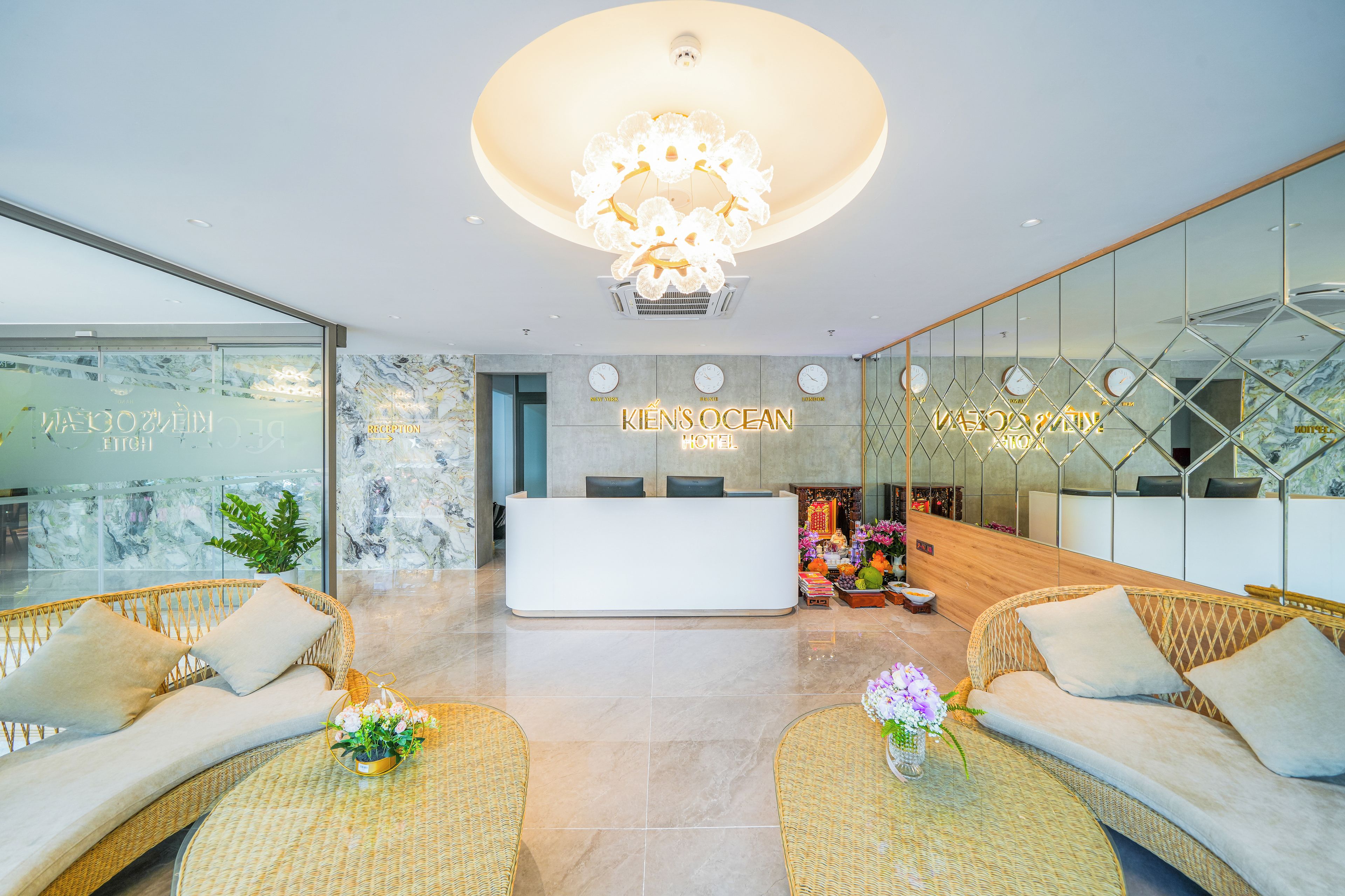 A hotel reception area with a modern design, featuring a reception desk, seating area with wicker furniture, and a large chandelier.