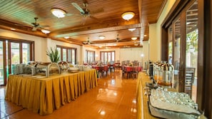 Un comedor espacioso con una larga mesa de buffet, varios ventiladores de techo y amplias ventanas que dejan entrar luz natural.