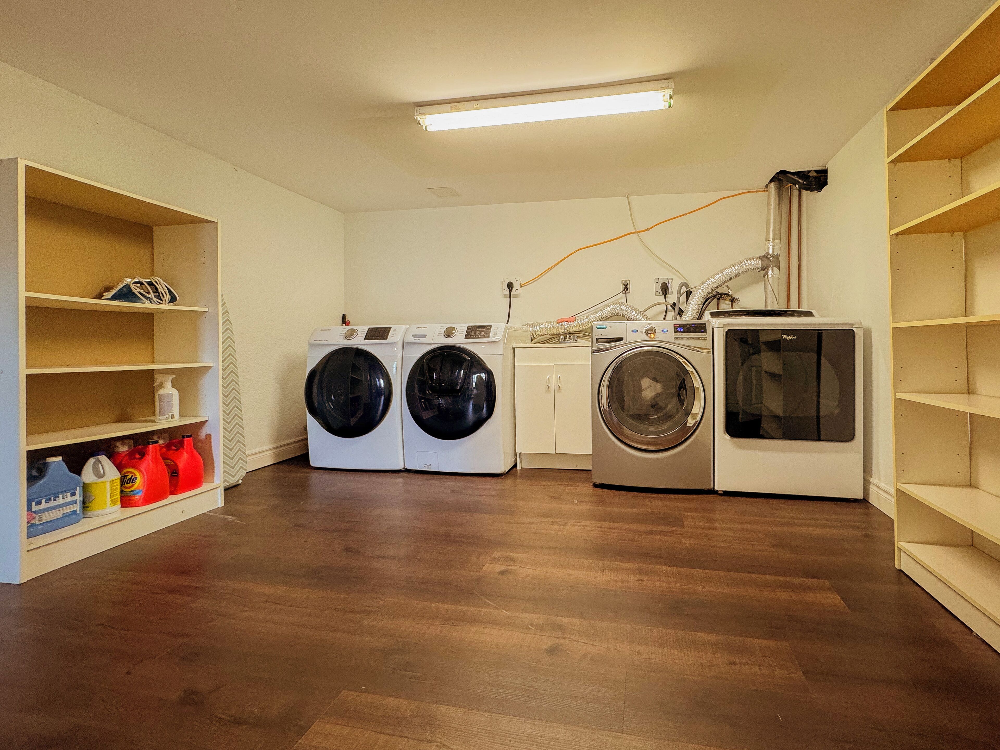 A laundry room with two front-loading washers and a dryer, wooden flooring, and shelves with laundry supplies.