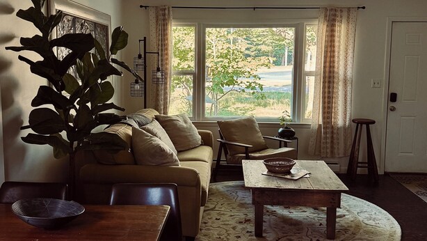 A living room with a sofa, coffee table, and dining table. There is a large window with curtains, a plant, and a collection of vintage soda bottles on the wall. - Chic Dunes Cottage W/ HOT TUB near Warren Dunes / Lake MI! (Shorewood-Tower Hills-Harbert)