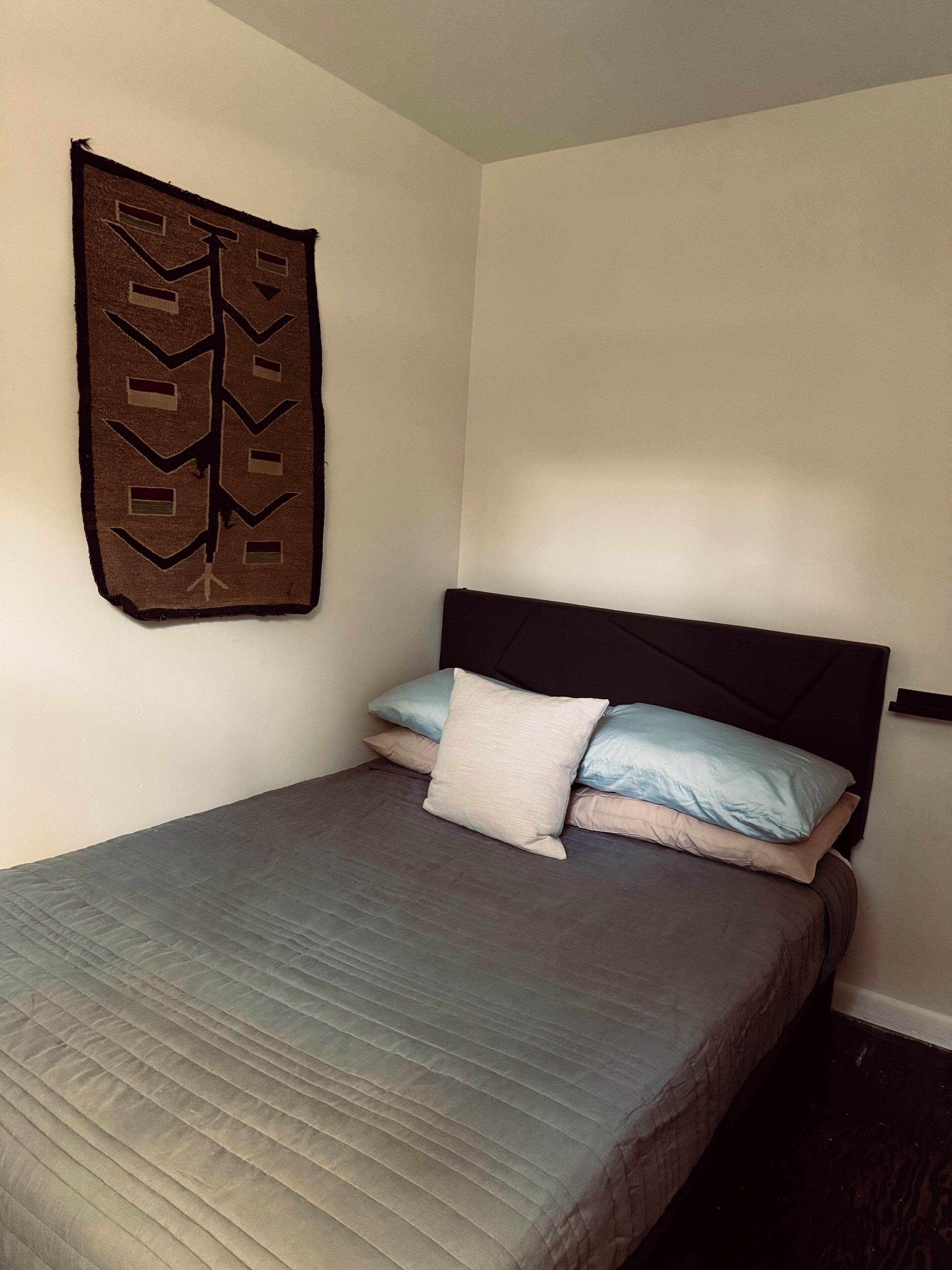 A single bed with a dark headboard, a quilted grey bedspread, and three pillows in shades of blue and beige. A patterned wall hanging is visible above the bed.