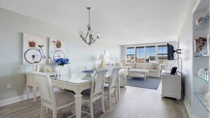 A spacious living area with a dining table, white furniture, and a large window with blinds. - Light bright airy loggerhead cay unit 314 New! (Sanibel)