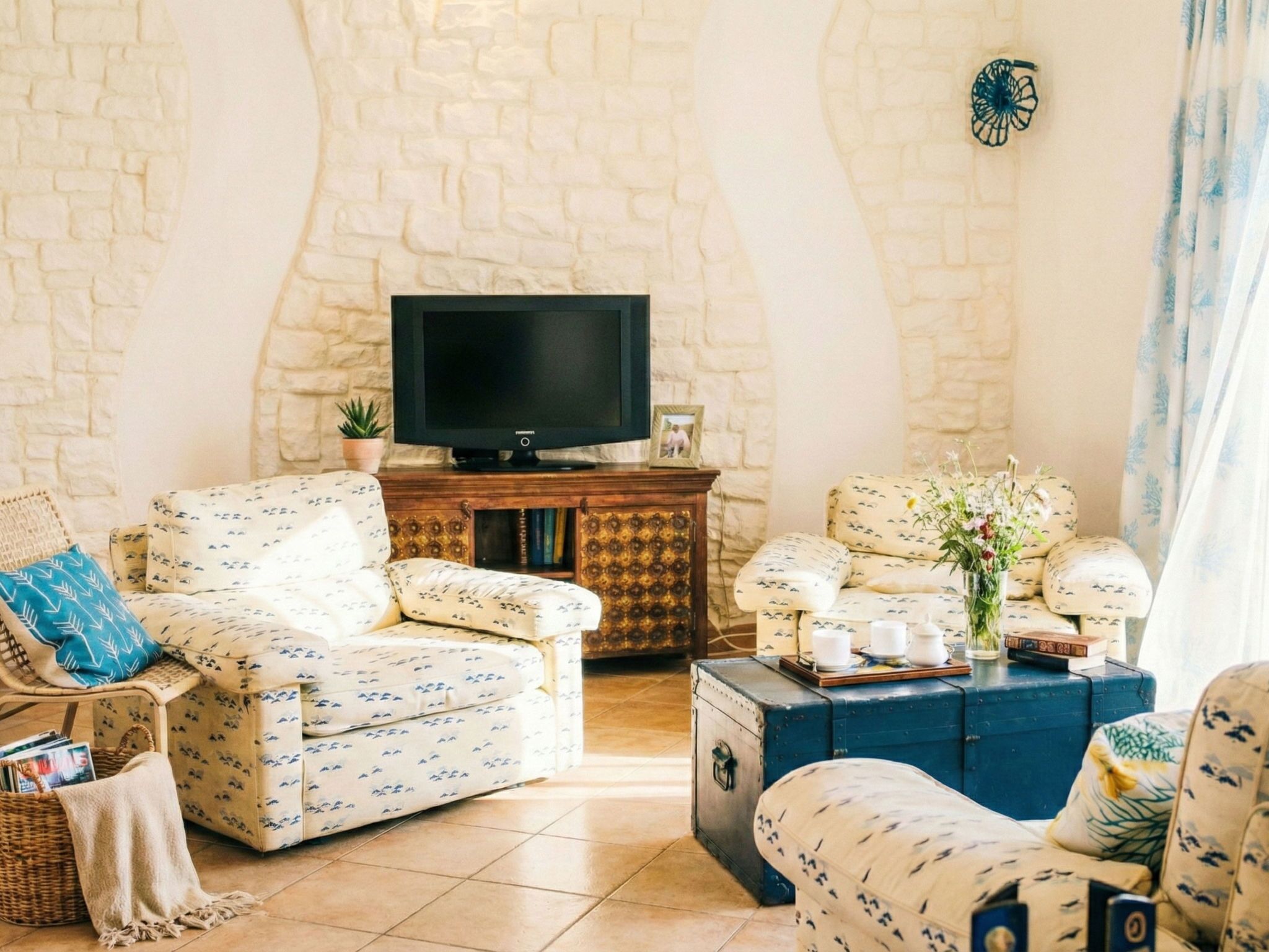 A cozy living room with a stone wall, a television, patterned sofas, a wooden cabinet, and a blue trunk.