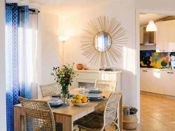A dining area with a wooden table set for four, surrounded by wicker chairs. A vase with flowers and a bowl of fruit are on the table.