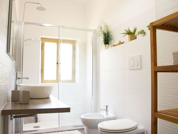 A modern bathroom with a large mirror, a sink, a toilet, and a shower. There is a window providing natural light and a shelf with plants and a teddy bear.