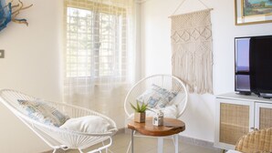 Une pièce dotée d’une grande fenêtre, d’une décoration murale en macramé, d’un téléviseur et de deux fauteuils en osier.