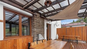 Une terrasse en bois avec une grande table et des bancs, un mur en pierre et un espace abrité doté d’un ventilateur de plafond.