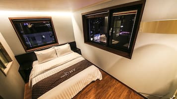 A hotel room with a bed, two windows, a wooden floor, and a night view of the city.