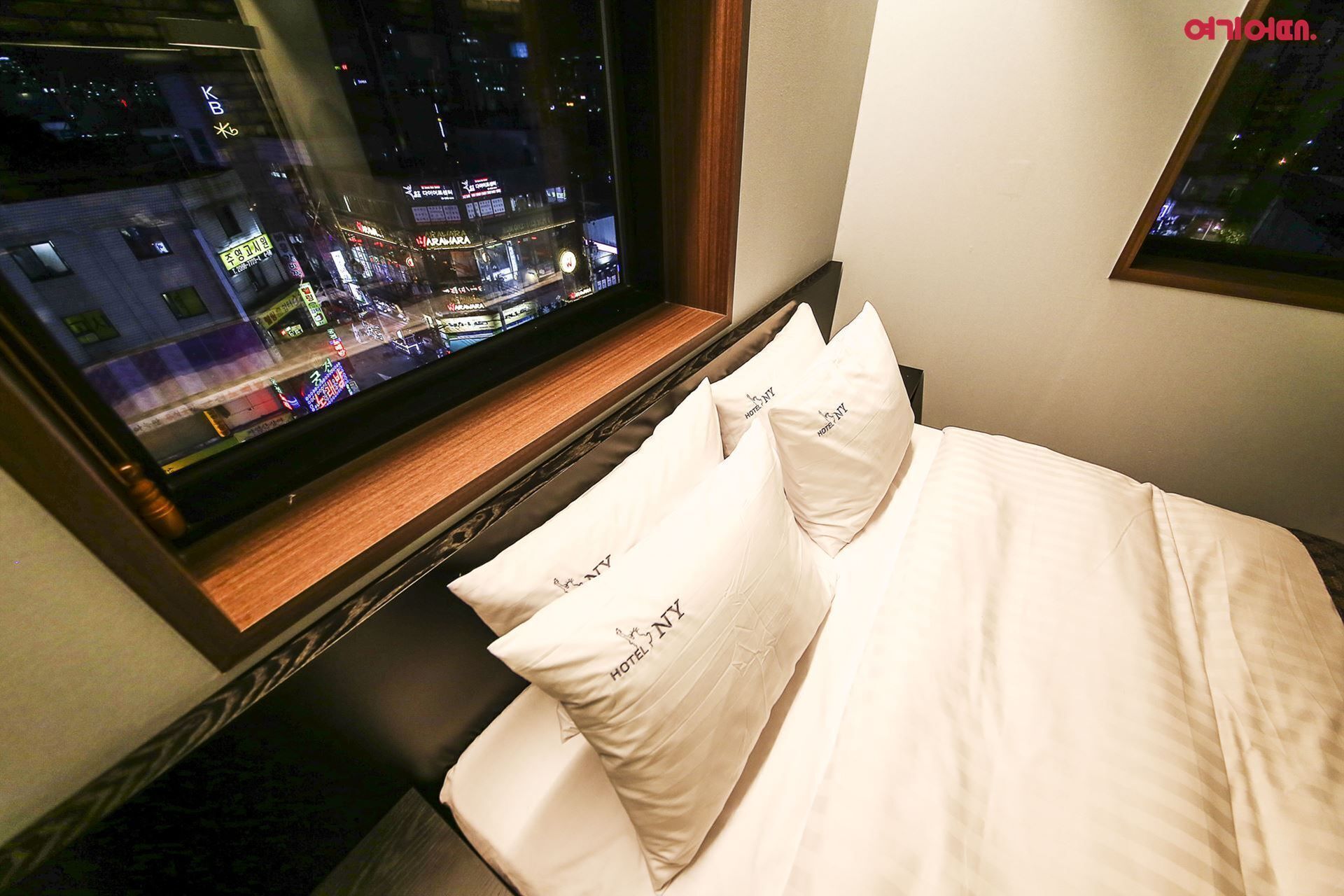 Una camera d'albergo con un letto, cuscini e una finestra che si affaccia su una vista notturna della città.