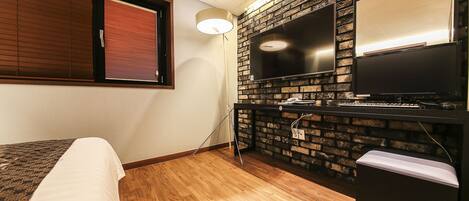 Habitación de hotel con pared de ladrillo, televisor de pantalla plana, escritorio con computadora, cama con cubrecama estampado y ventana con persianas.