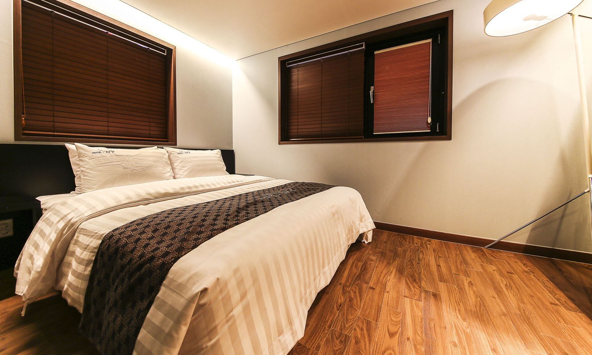 A bedroom with a bed, wooden flooring, and two windows with blinds.
