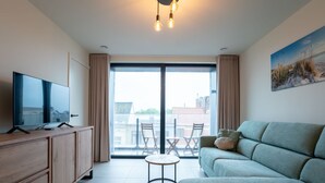A modern living room with a flat-screen TV on a wooden cabinet, a green sofa, a small round table, and a view of the city through large sliding doors. - Elyana with balcony by Interhome (Blankenberge)