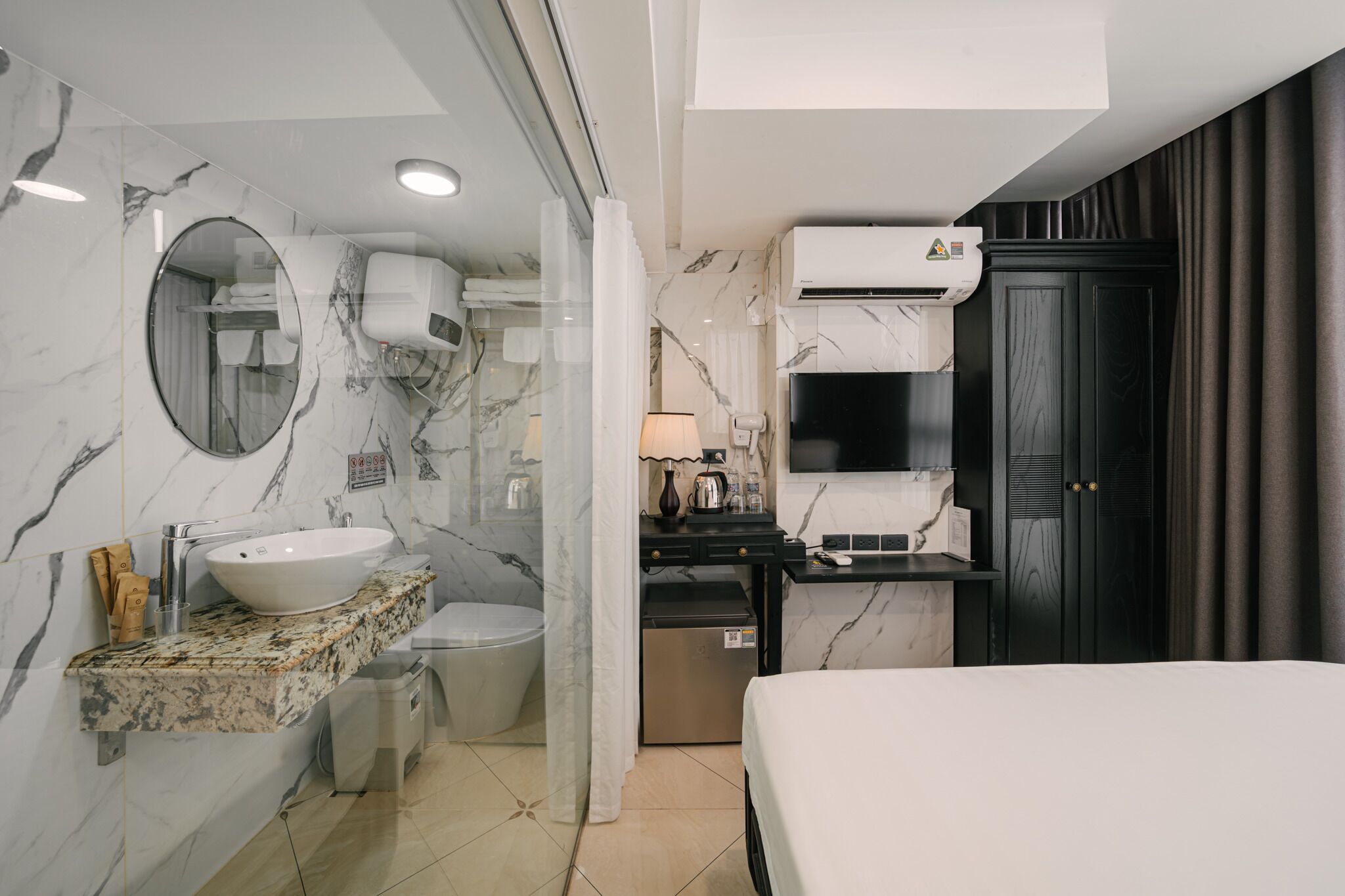 Un baño moderno con lavamanos, inodoro y zona de ducha de mármol, un pequeño escritorio con lámpara, y una cama.