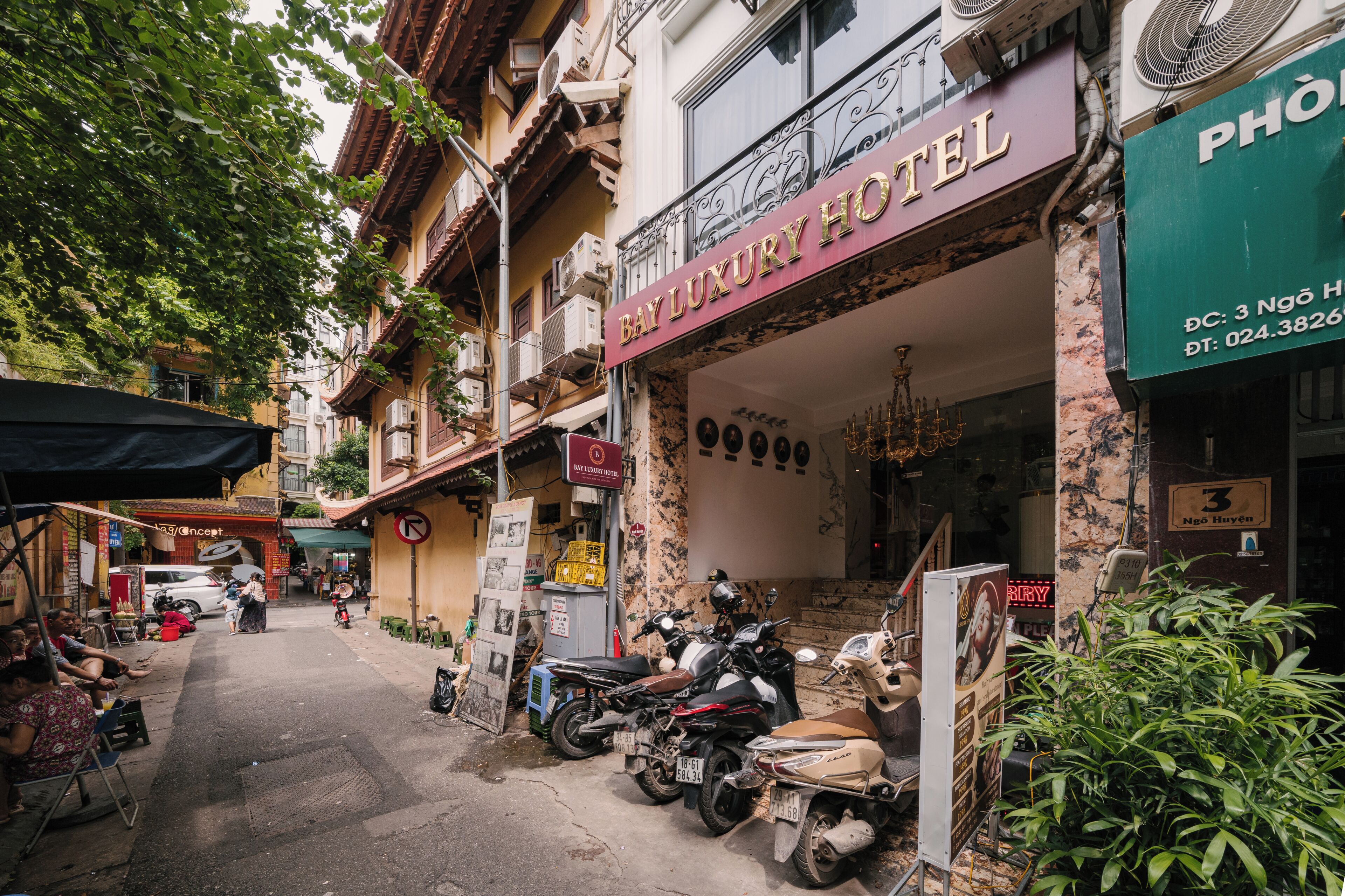 Vista da rua com um hotel de luxo chamado 'Bay Luxury Hotel' à direita. Motocicletas estão estacionadas na frente e um vendedor ambulante com um carrinho à esquerda.