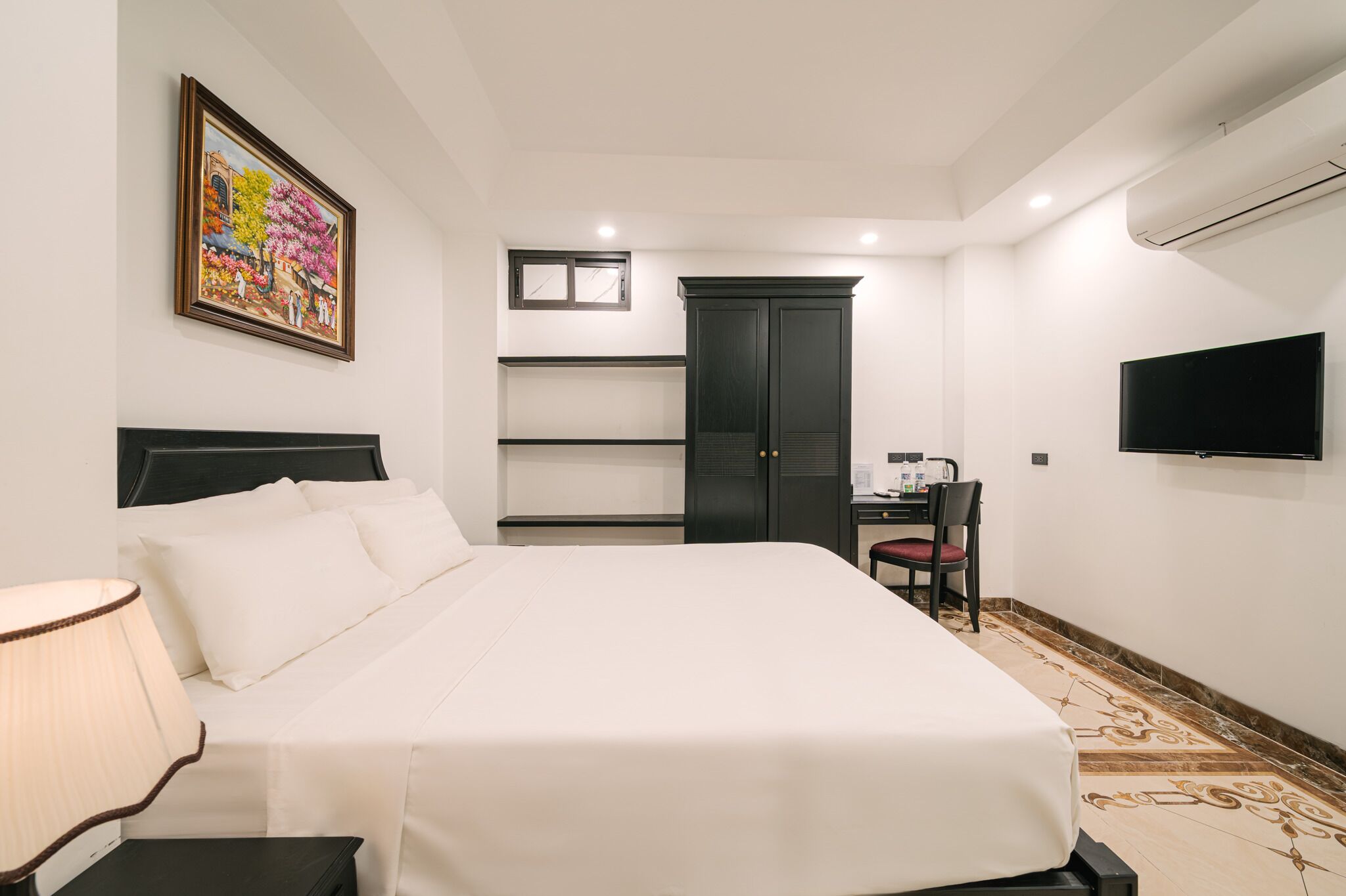 A hotel room with a bed, a desk, a chair, a TV, and a painting on the wall.