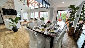Een moderne eetruimte met een lange tafel voor zes personen, omringd door witte stoelen. In het midden staat een grote potplant, een houten salontafel en een aan de muur bevestigde televisie. De kamer heeft een houten vloer en grote ramen voor natuurlijk licht.
