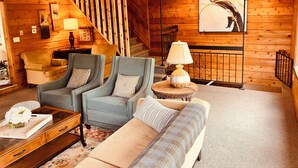 A cozy living room with a sofa, two armchairs, a coffee table, and a lamp. There is a staircase in the background and a painting on the wall. - >>The Daydream Lodge<<
@Racers Ridge (Plymouth)