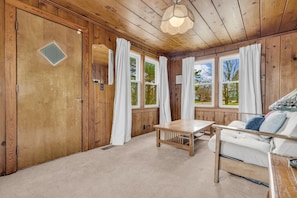 A cozy room with wooden walls, a large window with curtains, a wooden table, and a sofa with cushions.