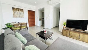 A modern living room with a sofa set, a coffee table, a TV on a wooden stand, and a dining area with chairs and a table. - Elysia Park B7- Luxury 2BR with Private Balcony & Resort Pools, gym-Free parking (Paphos)