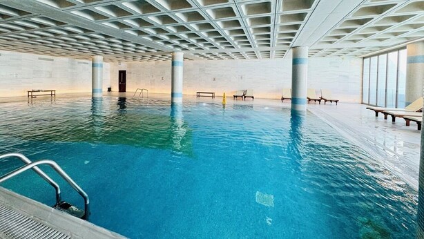 An indoor swimming pool with clear blue water, a ceiling with geometric patterns, and lounge chairs along the sides. - Elysia Park B7- Luxury 2BR with Private Balcony & Resort Pools, gym-Free parking (Paphos)