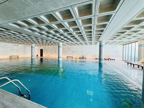 An indoor swimming pool with clear blue water, a ceiling with geometric patterns, and lounge chairs along the sides. - Sunny 2BR at Elysia Park w/ Balcony, Outdoor & Heated Pool, Gym - Central Paphos (Paphos)