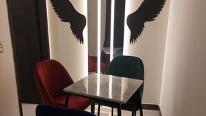 Una habitación pequeña con una mesa y tres sillas, una roja y dos azules, y una pared decorada con dos grandes siluetas negras de alas.
