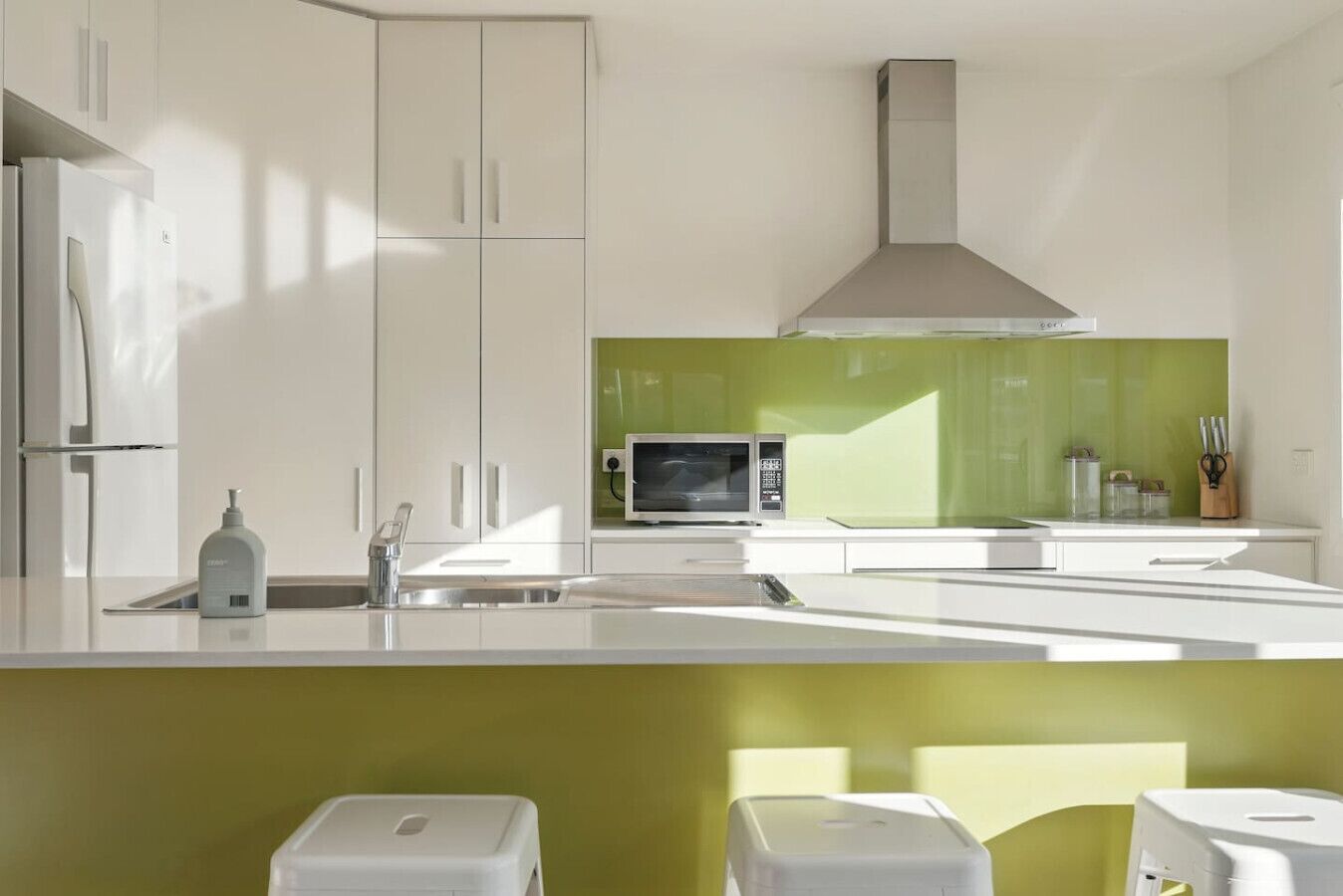 Una cuina moderna amb armaris blancs, un frontissos verd i un microones integrat, i una campana extractora d'acer inoxidable.