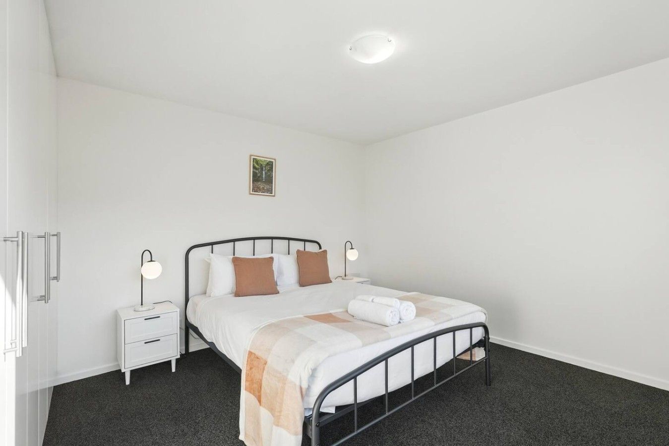 Ein Einzelbett mit Metallgestell, ein Nachttisch mit Lampe und ein kleines Bild an der Wand.