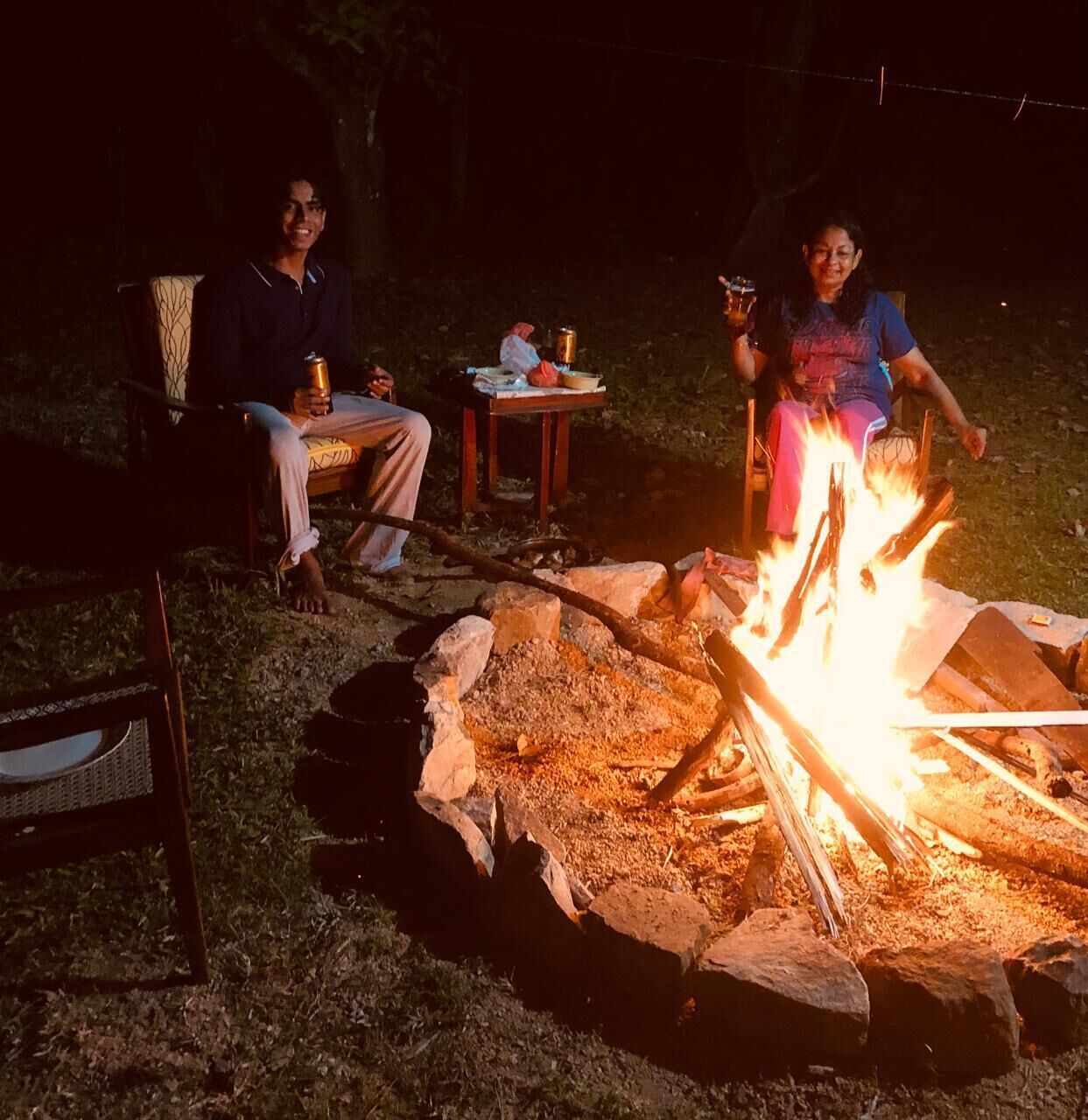 An outdoor setting with a campfire, two people sitting on chairs, and a table with items.
