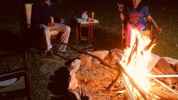 An outdoor setting with a campfire, two people sitting on chairs, and a table with items.
