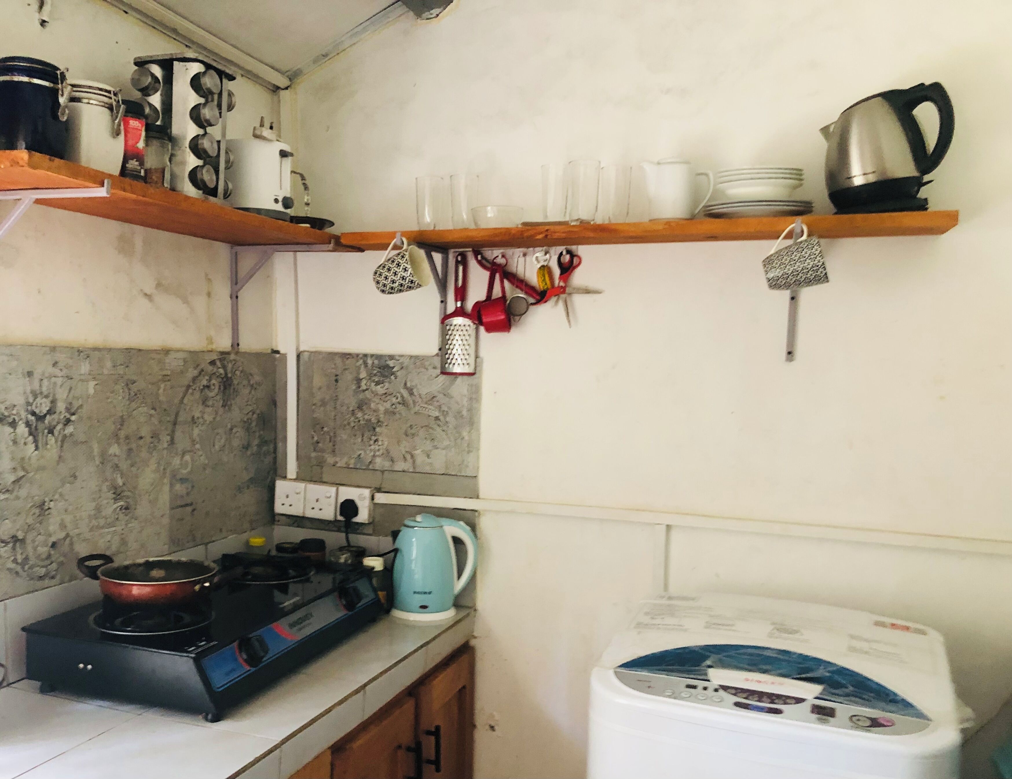 Een keuken met een gasfornuis, waterkoker en een wasmachine. Er zijn planken met servies en een rek met keukengerei.
