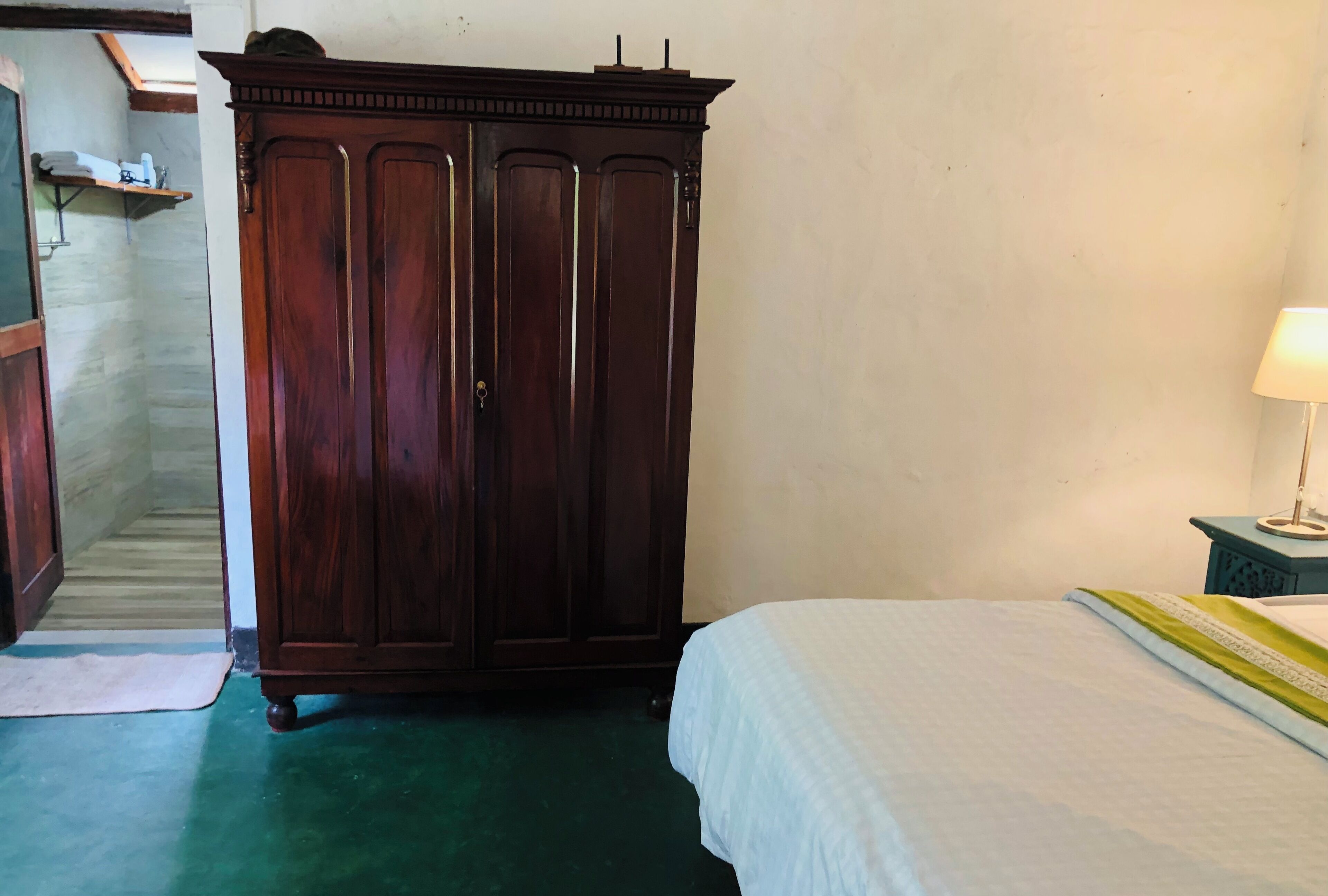 A room with a bed, a wooden wardrobe, and a small shelf with a lamp.