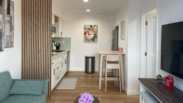 Een moderne woonkamer met een bank, een salontafel met bloemen, een televisie en een keukenhoek met kastjes en een magnetron.