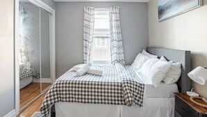 Um quarto com colcha xadrez, luminária de cabeceira, espelho e janela com cortinas.