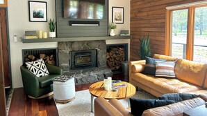 A cozy living room with a stone fireplace, leather sofas, a flat-screen TV, and a large window offering a view of the outdoors.