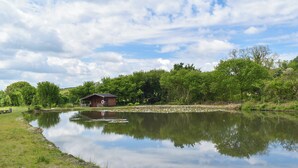 A tranquil lakeside scene with a wooden cabin, lush greenery, and a clear sky.