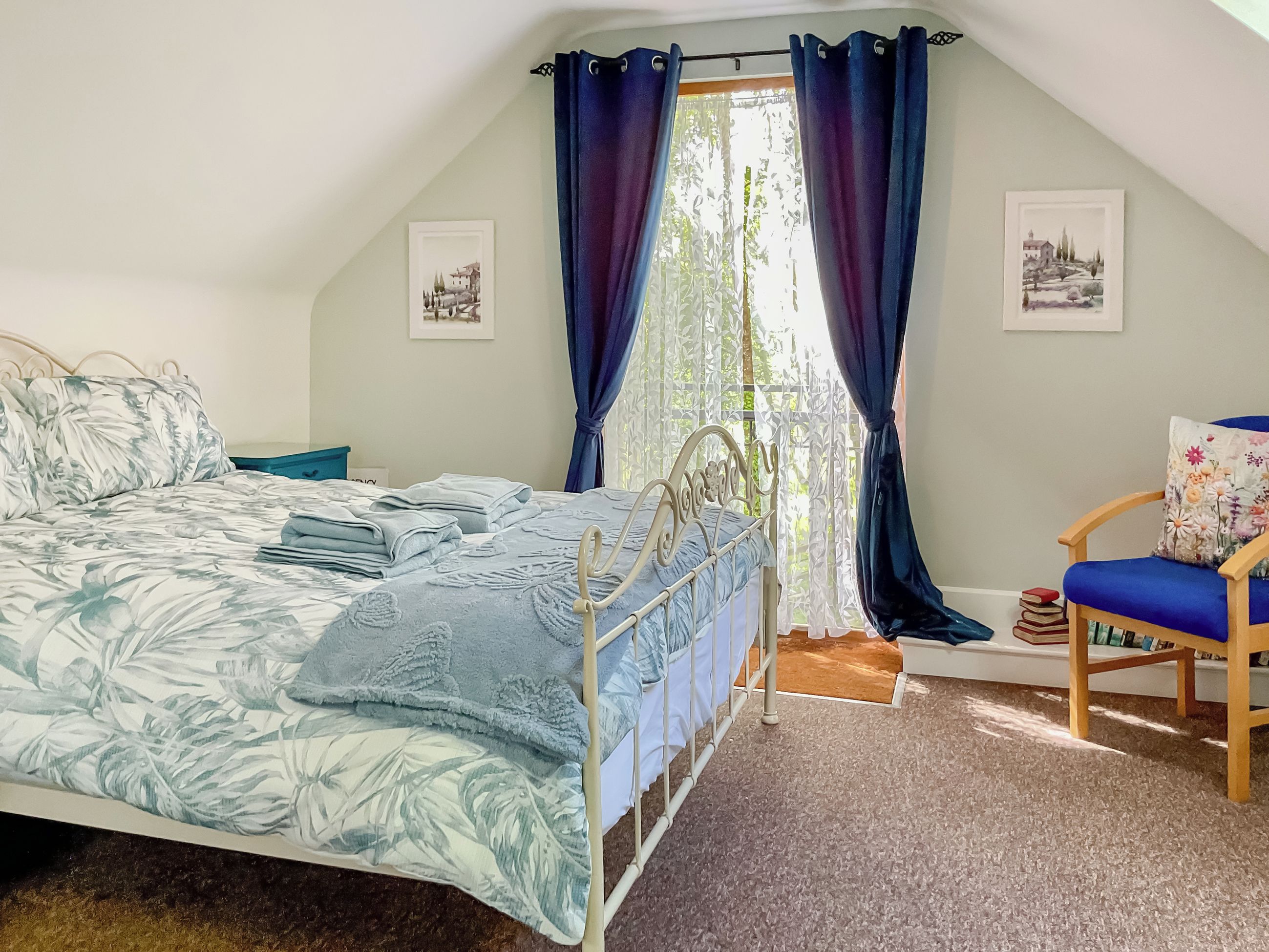 A bedroom with a bed, a chair, a window with curtains, and two framed pictures on the wall.