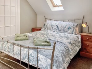 A bed with a metal frame, a floral bedspread, two green towels, a wooden nightstand with a lamp, and a skylight above.