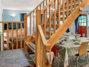 Un escalier en bois mène à un coin repas où une table est dressée pour deux personnes. On aperçoit une télévision au fond et, par une fenêtre, on a une vue sur l’extérieur.